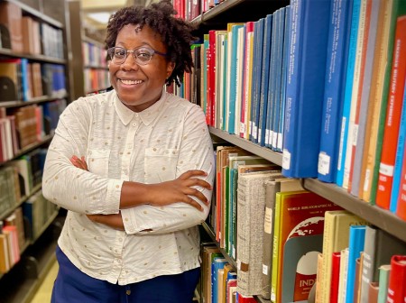 Moya Bailey stands smiling with her arms crossed next to large shelves of books at a library.