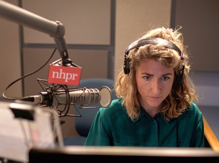 Lauren Chooljian wears headphones while sitting at a desk in a radio studio. There is a microphone at her right labeled “NHPR.”