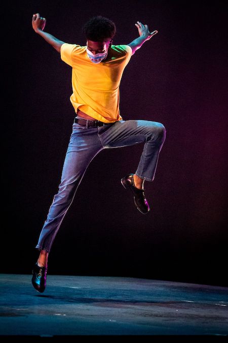 Sterling Harris tap dancing on a stage while wearing a mask. He is pictured mid-jump, with his left knee bent and high up off the ground, while his right foot has just barely left the ground. His arms are extended behind him.