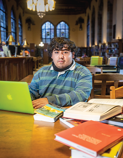 Daniel Rodriguez sits at a desk inside a library, with his laptop and several open books scattered in front of him. He wears a striped blue and green shirt. 