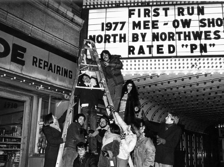 Members of the 1977 Mee-Ow Show cast pose in front of the marquee at Evanston’s Varsity Theater, photo in black and white.