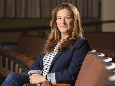 Ana Gasteyer sits in a row of auditorium chairs, smiling with her legs crossed.