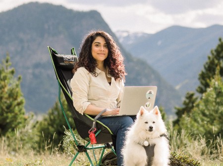 Alexa Carreno sits in a camping chair with a laptop on her lap and a furry white dog by her side, in front of large mountains. 