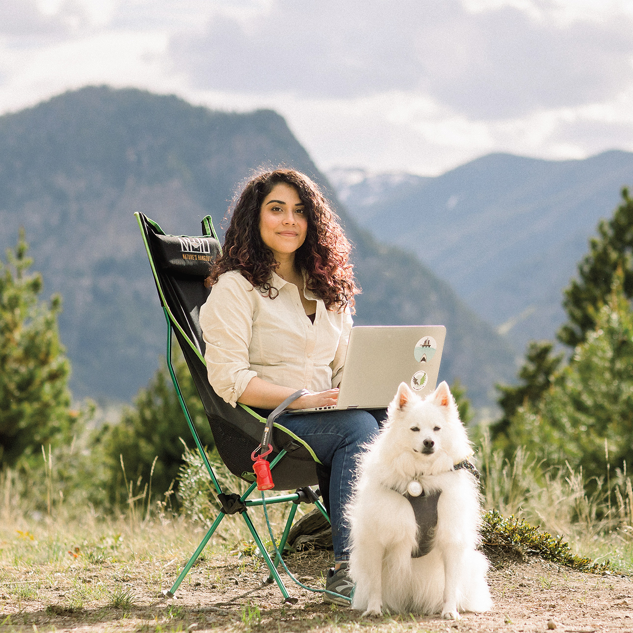 Alexa Carreno sits in a camping chair with a laptop on her lap and a furry white dog by her side, in front of large mountains. 