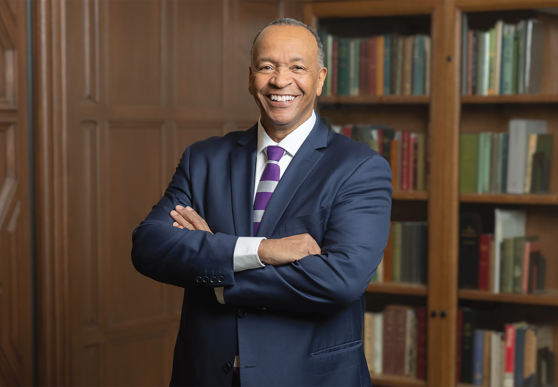 Charles Modlin stands tall, smiling with his arms crossed in front of a bookshelf.