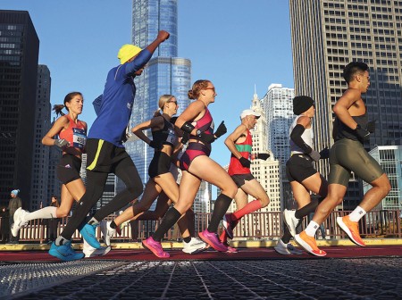 Chicago Marathon runners stride over a bridge that crosses over the river, with tall skyscrapers in the back.