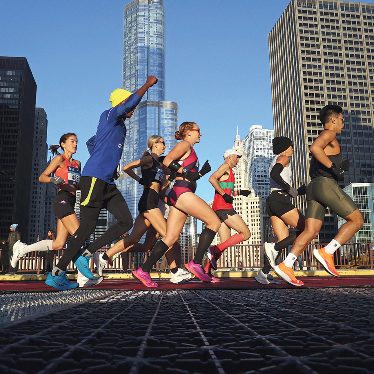 Chicago Marathon runners stride over a bridge that crosses over the river, with tall skyscrapers in the back.