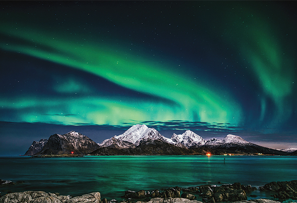 Mountains and the ocean sit below a large display of northern lights.