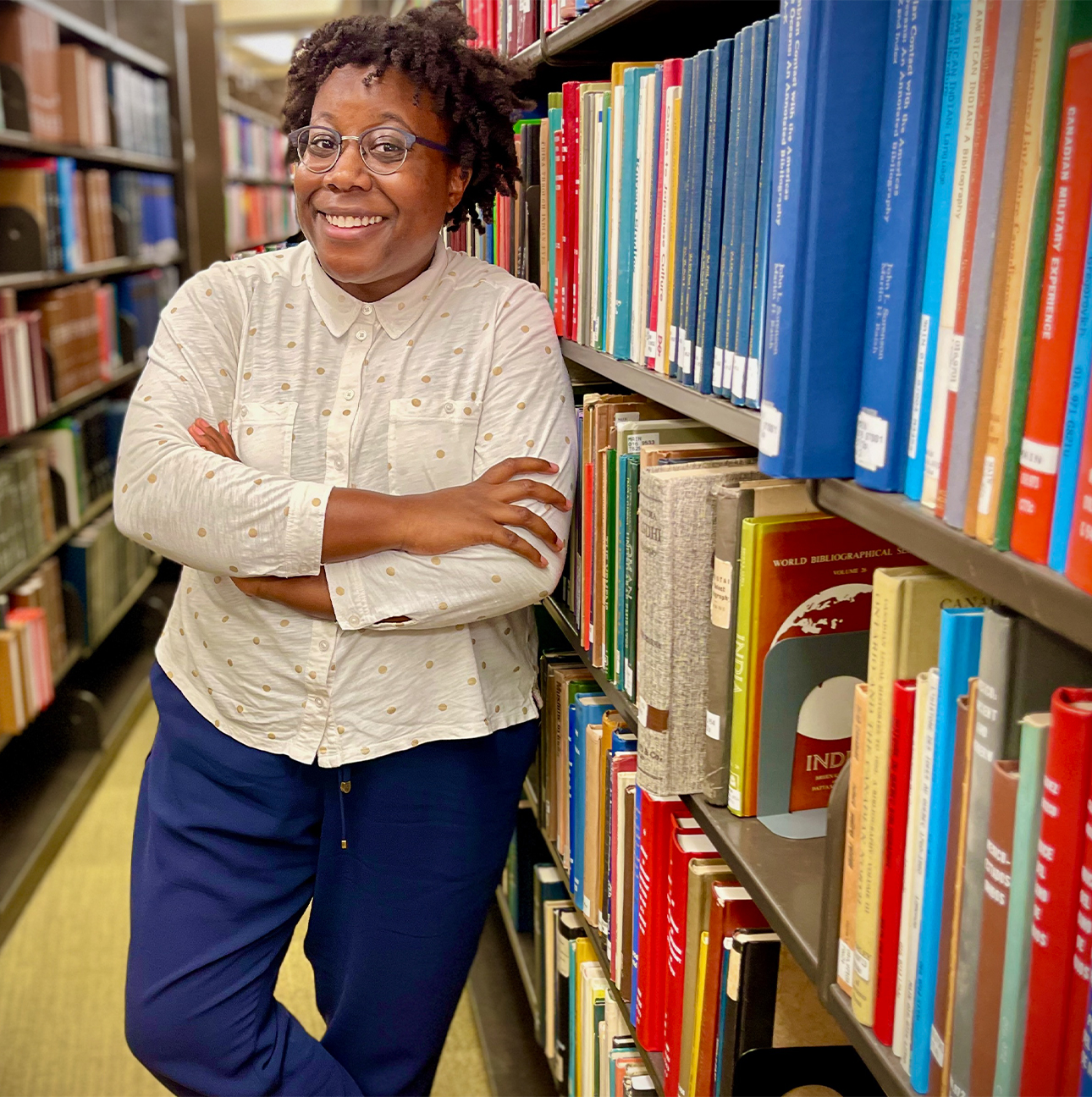 Moya Bailey stands smiling with her arms crossed next to large shelves of books at a library.