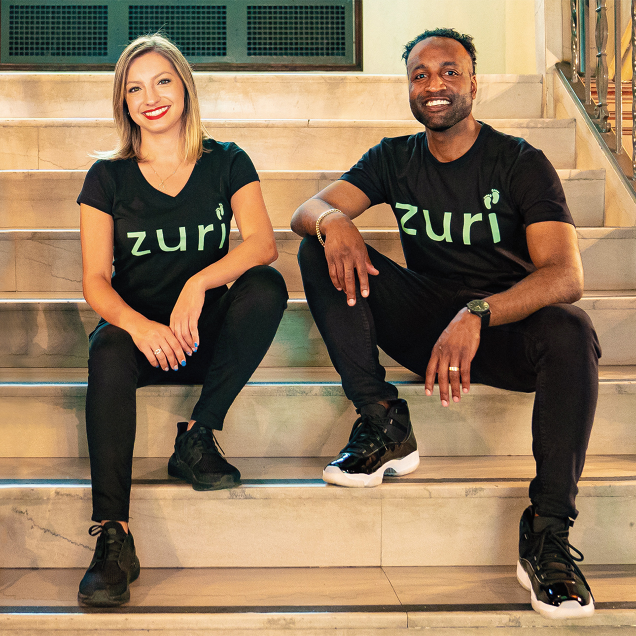 Giuliana Zaccardelli and Blair Matthews sit on steps next to each other smiling for a photo, both are wearing "Zuri" shirts.