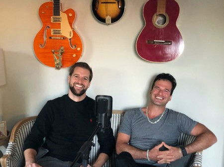 Musical duo Kevin Hoban and Jordan Simkovic sit in front of a podcast mic smiling, with their guitars behind them.