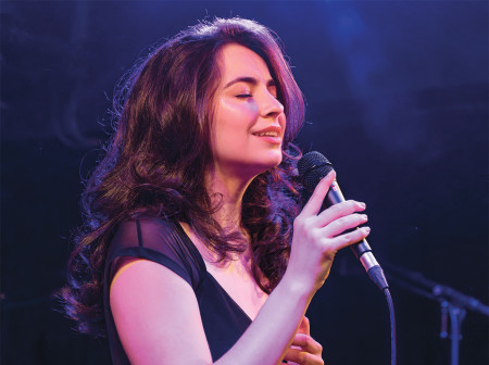 Stella Cole sings with her eyes closed while holding a microphone. She is onstage in London. She is wearing a black dress.