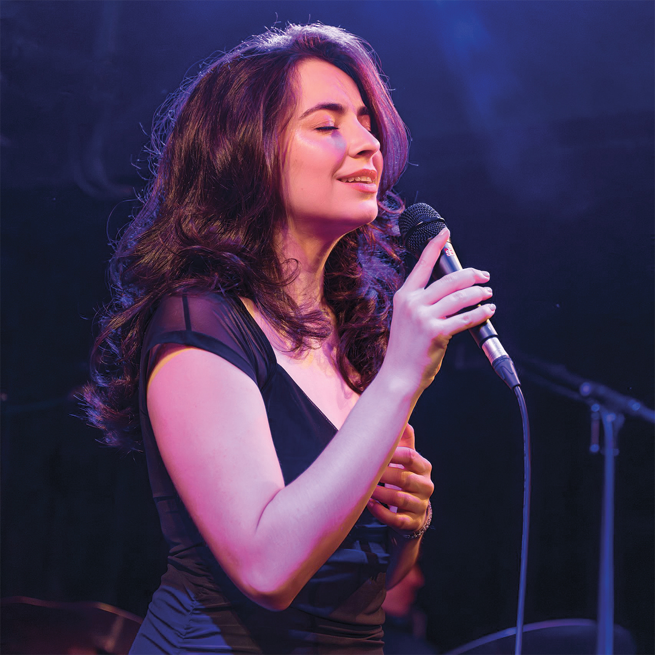 Stella Cole sings with her eyes closed while holding a microphone. She is onstage in London. She is wearing a black dress.