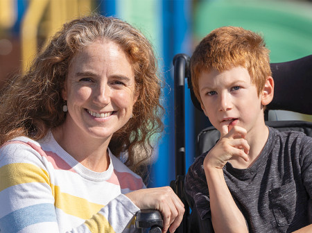 Annah Abetti Korpi smiles into the camera as she crouches next to her son, Isidoro Korpi, who sits in a wheelchair. There is a playground in the background. Annah is wearing a gray shirt with pink, red, blue and yellow stripes and Isidoro is wearing a gray T-shirt and blue pants. 