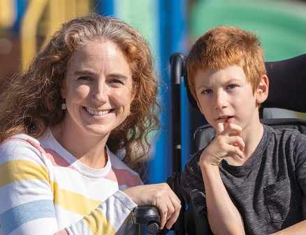 Annah Abetti Korpi smiles into the camera as she crouches next to her son, Isidoro Korpi, who sits in a wheelchair. There is a playground in the background. Annah is wearing a gray shirt with pink, red, blue and yellow stripes and Isidoro is wearing a gray T-shirt and blue pants. 