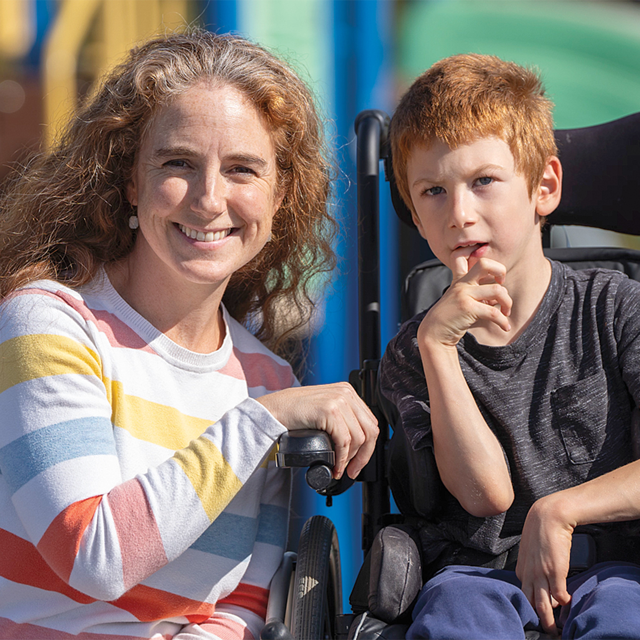 Annah Abetti Korpi smiles into the camera as she crouches next to her son, Isidoro Korpi, who sits in a wheelchair. There is a playground in the background. Annah is wearing a gray shirt with pink, red, blue and yellow stripes and Isidoro is wearing a gray T-shirt and blue pants. 