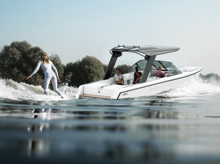 Three people sit aboard the Arc Sport EV boat. A woman wake boarder in a silver suit rides on a board behind the boat. Her back is to the viewer. There are green trees in the background.