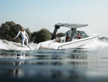 Three people sit aboard the Arc Sport EV boat. A woman wake boarder in a silver suit rides on a board behind the boat. Her back is to the viewer. There are green trees in the background.