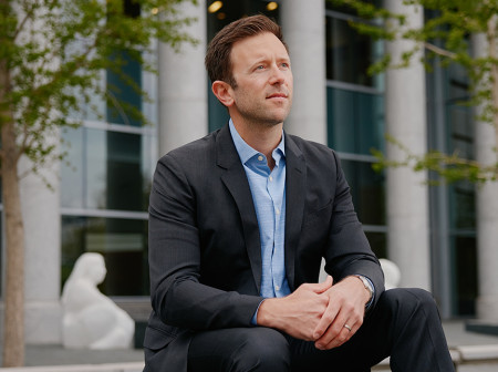 Beau Tremitiere sits on a ledge in front of a cement building with his arms resting on his thighs and his hands clasped as he looks off into the distance. He is wearing a black suit with a light blue dress shirt.