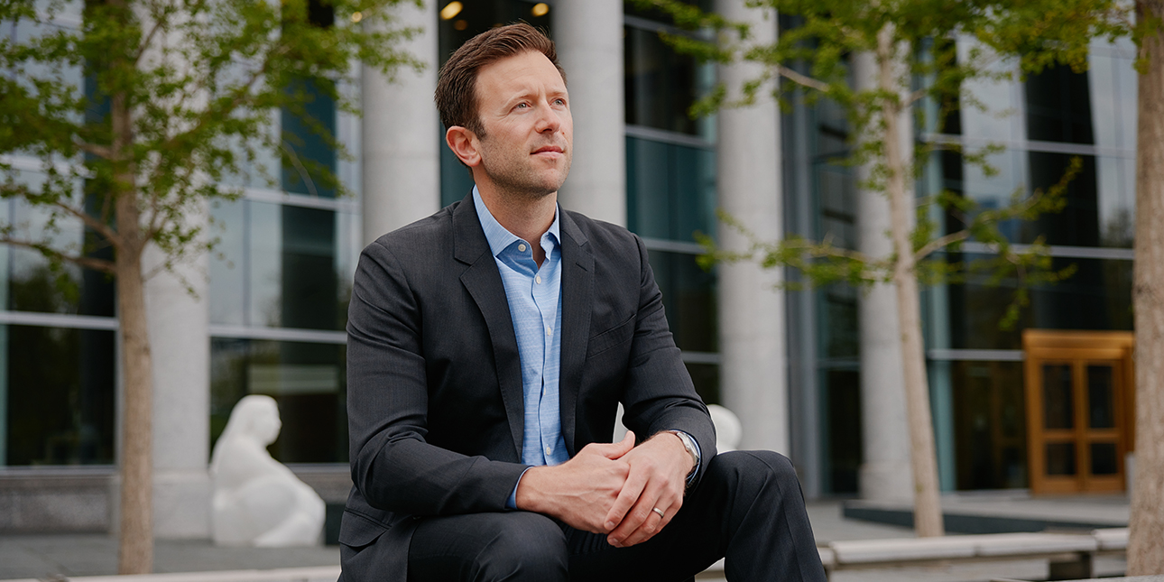 Beau Tremitiere sits on a ledge in front of a cement building with his arms resting on his thighs and his hands clasped as he looks off into the distance. He is wearing a black suit with a light blue dress shirt.