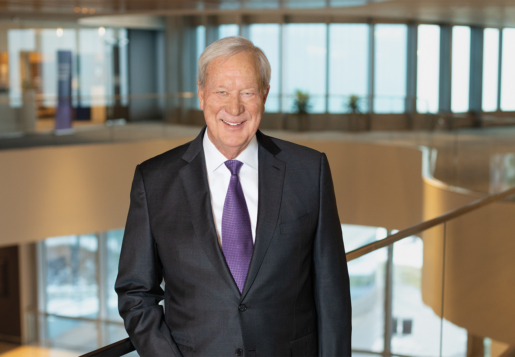 Bill Osborn stands in a spacious, windowed hallway with one hand in his pocket, wearing a black suit and purple tie and smiling at the camera.