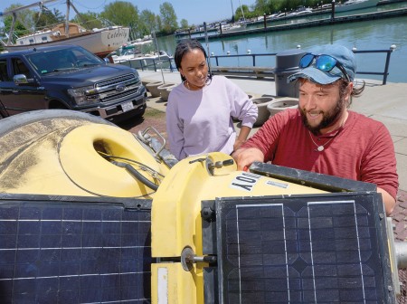 Ben Szczygiel holds a large yellow buoy fitted with solar panels on its side while Sydney Williams observes. There is a waterway in the background, with a truck and boat parked nearby. Szczygiel is wearing a red long sleeve shirt, blue baseball cap and sunglasses, and Williams is wearing a lavender sweatshirt.