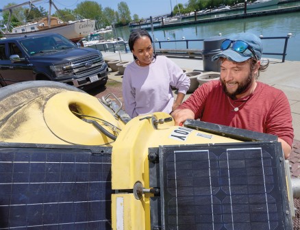 Ben Szczygiel holds a large yellow buoy fitted with solar panels on its side while Sydney Williams observes. There is a waterway in the background, with a truck and boat parked nearby. Szczygiel is wearing a red long sleeve shirt, blue baseball cap and sunglasses, and Williams is wearing a lavender sweatshirt.