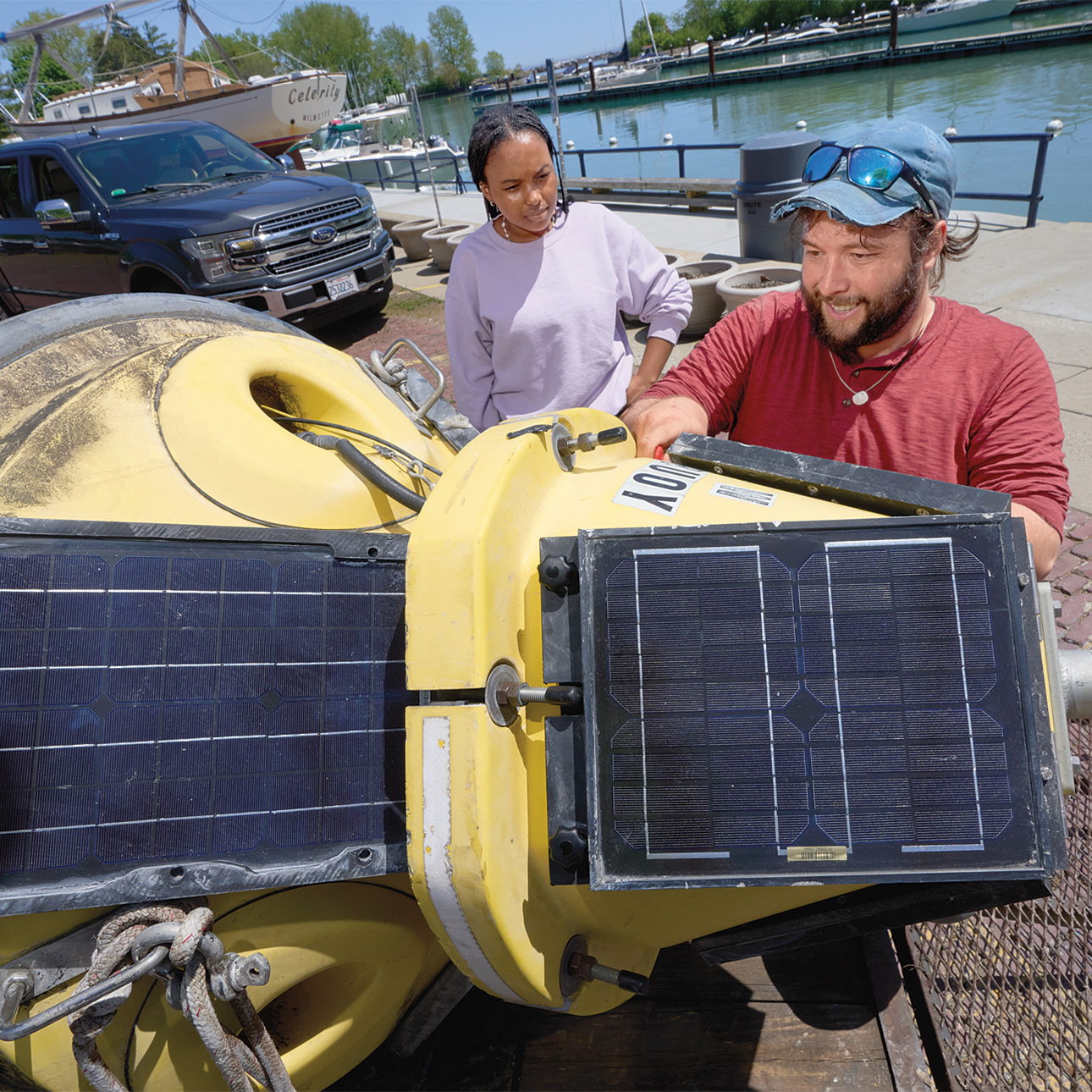 Ben Szczygiel holds a large yellow buoy fitted with solar panels on its side while Sydney Williams observes. There is a waterway in the background, with a truck and boat parked nearby. Szczygiel is wearing a red long sleeve shirt, blue baseball cap and sunglasses, and Williams is wearing a lavender sweatshirt.