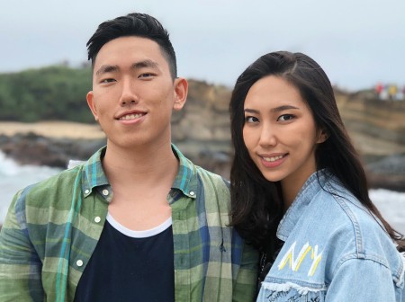 Ryan and Chelsea Jeon smile side-by-side in front of a rocky landscape. Ryan is wearing a black shirt and green flannel button-down, and Chelsea is wearing a denim jacket with painted designs.