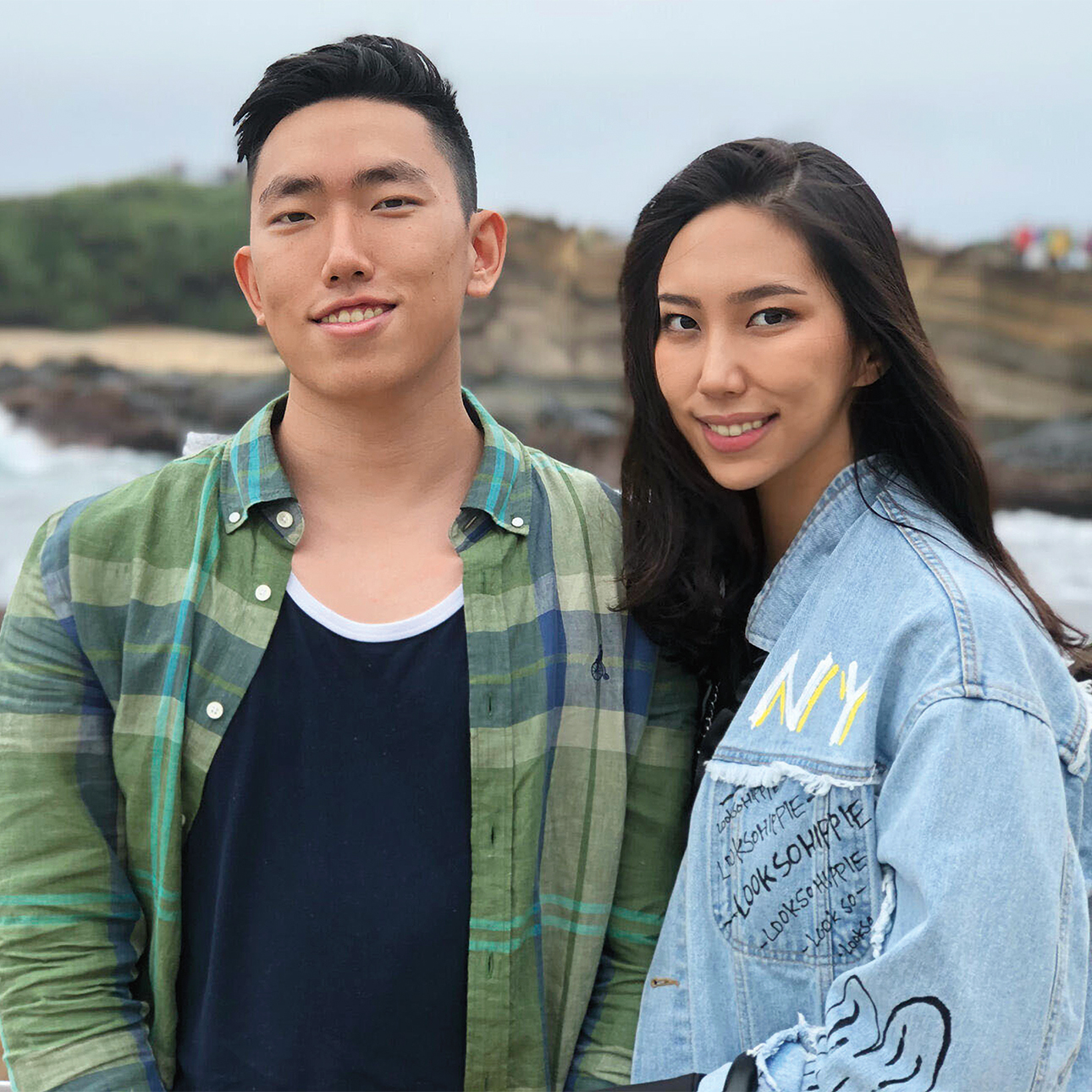 Ryan and Chelsea Jeon smile side-by-side in front of a rocky landscape. Ryan is wearing a black shirt and green flannel button-down, and Chelsea is wearing a denim jacket with painted designs.