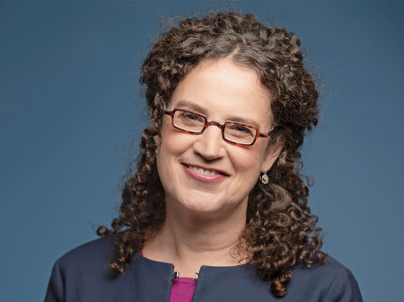 Deborah Cohen smiles at the camera in front of a blue background. She wears a purple shirt, a dark blue jacket and glasses.