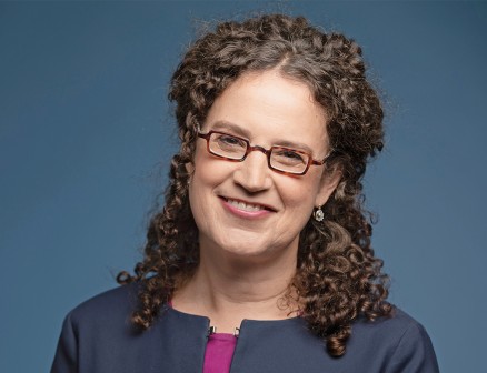 Deborah Cohen smiles at the camera in front of a blue background. She wears a purple shirt, a dark blue jacket and glasses.