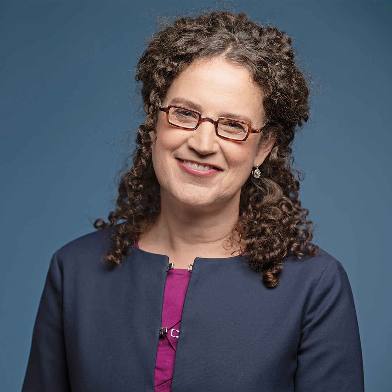 Deborah Cohen smiles at the camera in front of a blue background. She wears a purple shirt, a dark blue jacket and glasses.