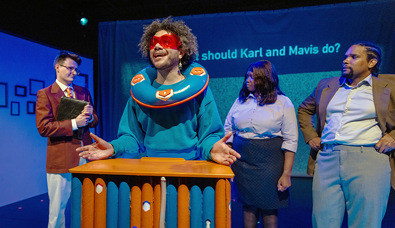 The character 4D, wearing a blue jumpsuit and orange glasses, stands at an orange and blue–colored podium and asks the audience to help the other characters behind him make a decision about housing. Behind 4D are the characters Mr. Murphy and Dot’s parents, Mavis and Karl. Projected onto the backdrop of the stage is the sentence: What should Karl and Mavis do?