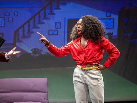 Two characters from the show Dot’s Home Live converse onstage. On the left is Mr. Murphy, a white man wearing a brown suit jacket and tan slacks and holding a black folder. On the left is Dot, a Black woman wearing a red top and white pants, pointing her finger at Mr. Murphy.