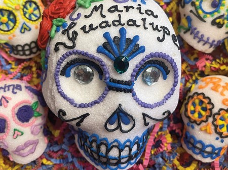 A box with five molded sugar skulls decorated with royal icing and jewels. The center skull is the largest and is decorated with royal blue and purple icing. The words Maria Guadalupe are written in black icing across the forehead. The surrounding skulls also have the names of lost loved ones written on their foreheads.