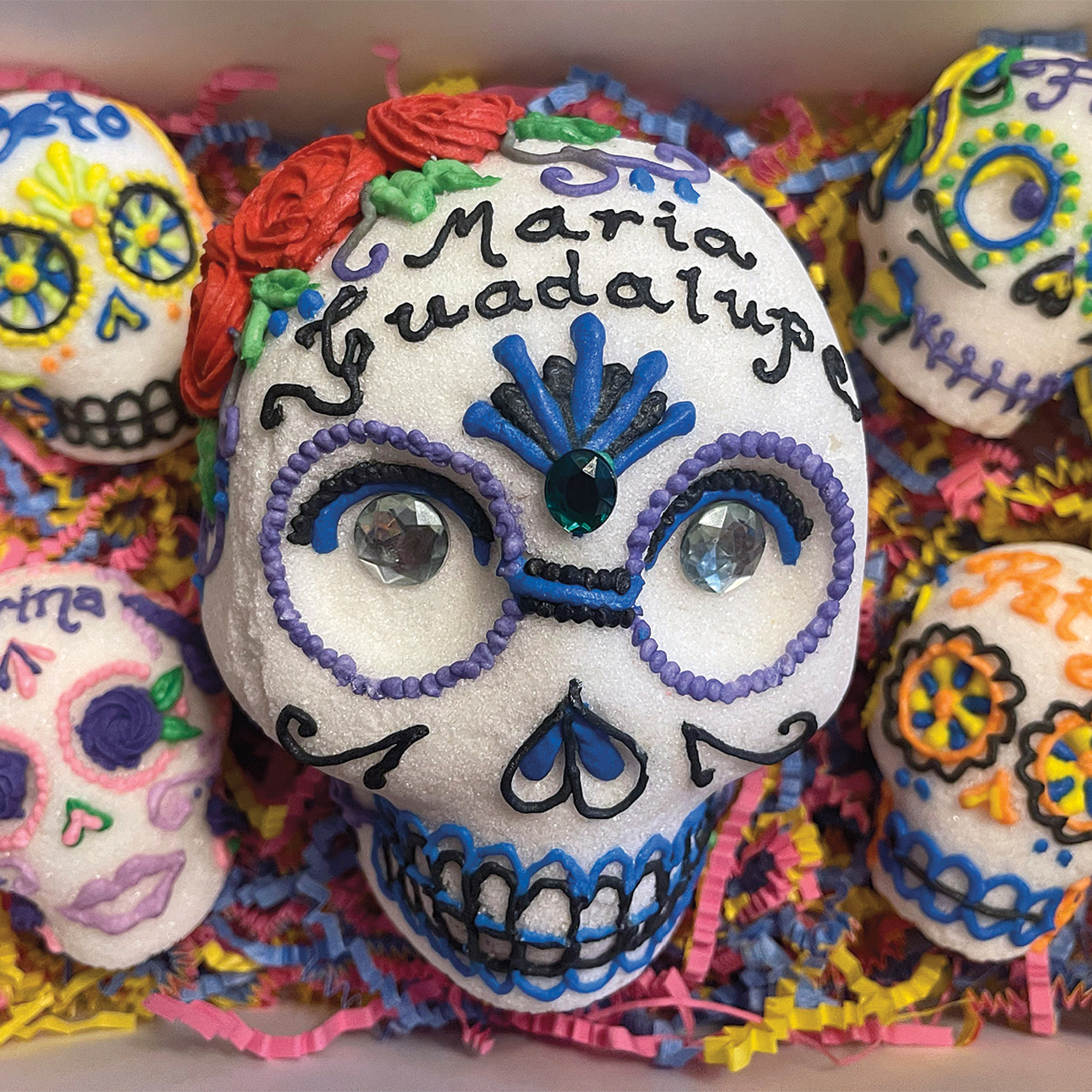 A box with five molded sugar skulls decorated with royal icing and jewels. The center skull is the largest and is decorated with royal blue and purple icing. The words Maria Guadalupe are written in black icing across the forehead. The surrounding skulls also have the names of lost loved ones written on their foreheads.