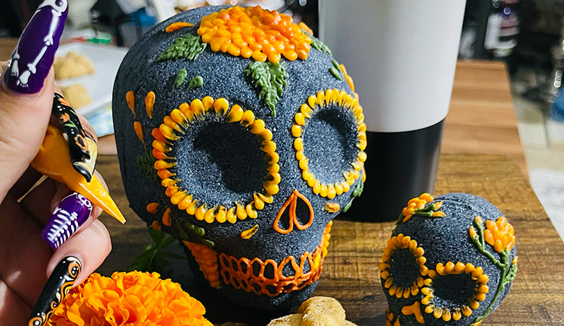 Two black sugar skulls decorated with orange and green flowers and a pan de muerto roll sit on a wooden table, alongside an orange flower and two icing tubes. 