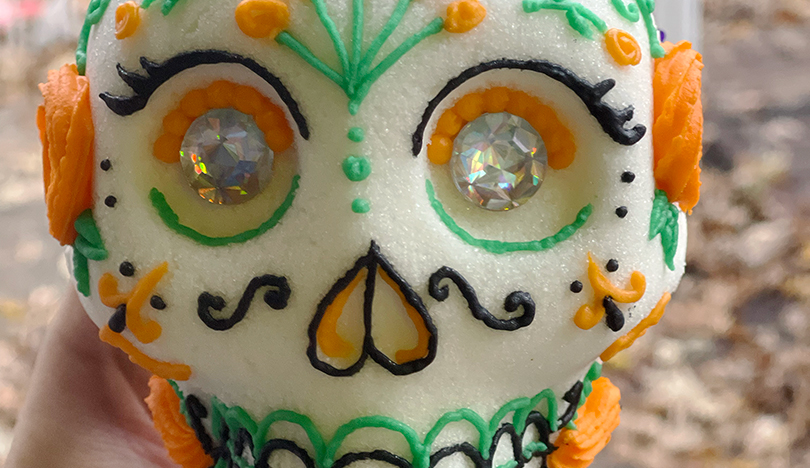 A white sugar skull decorated with black, orange and green icing in a floral pattern and jeweled eyes.