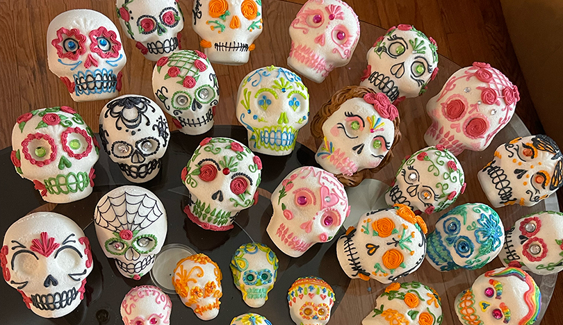 Several sugar skulls of varying sizes decorated with icing in various colors and patterns sit on a clear glass table over a wooden floor.