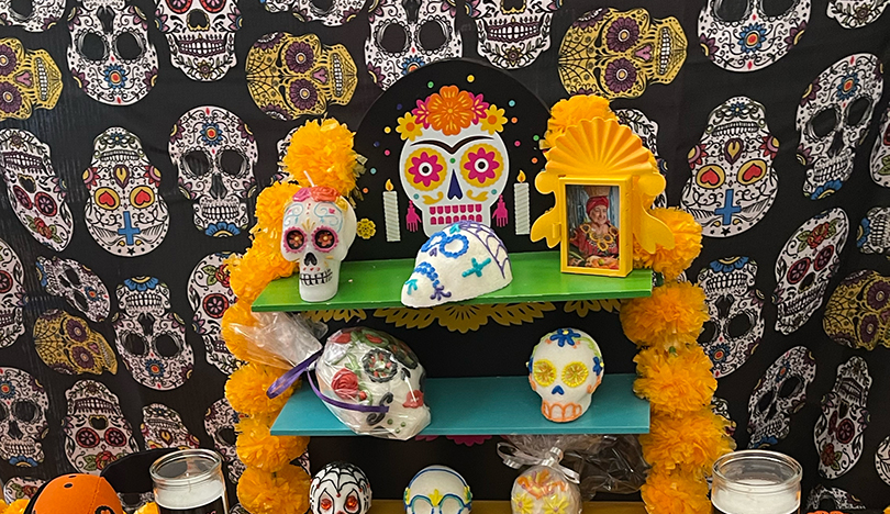 An ofrenda with a shelved display holds several decorated sugar skulls and a framed photo. There is a backdrop with a sugar skull pattern hanging behind the table. 