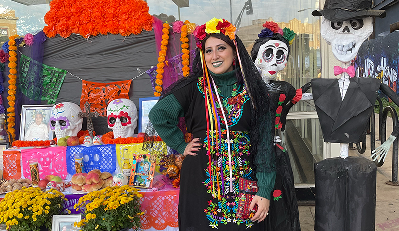 Genesis Garcia smiles with one hand on her hip beside a community altar. The altar is decorated with photographs, flowers, fruit, sugar skulls, candles and other offerings. Garcia is wearing a green turtleneck sweater, a black dress with colorful floral embroidery, black tights, brown boots and a yellow, orange and red flower crown. 