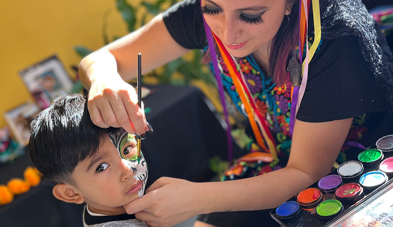 Genesis Garcia paints a child’s face with a skull design as he looks off-camera. In the foreground, a palette of face paints sits on a table with a black and orange tablecloth. Garcia is wearing a black dress with colorful floral embroidery and a yellow, orange and red flower crown.