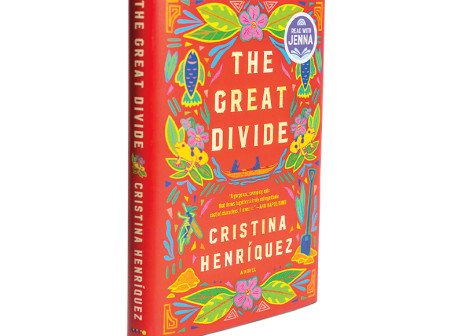 A photograph of The Great Divide book, which is angled slightly to the right. The book cover has a primarily red background with geometric green and yellow plants, flowers and frogs scattered around the title and the author’s name. 