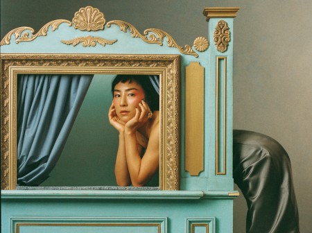 Greta Lee, bending at the waist, rests her elbows on a puppet theater with her head in the window behind the curtains. The puppet theater’s teal body is adorned with gold details, and Lee is dressed in a dark skirt with white heeled shoes.