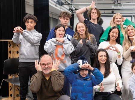 A group of children and adults pose in a mirrored studio room, holding up peace signs, hearts and thumbs up and smiling at the camera.