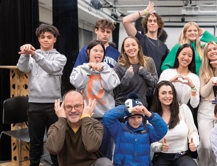 A group of children and adults pose in a mirrored studio room, holding up peace signs, hearts and thumbs up and smiling at the camera.