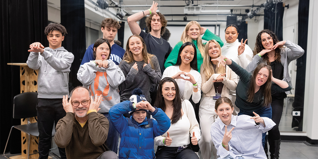 A group of children and adults pose in a mirrored studio room, holding up peace signs, hearts and thumbs up and smiling at the camera.