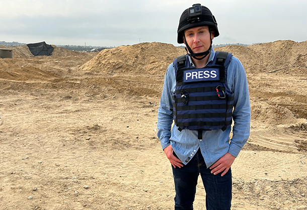 Josh Lederman stands in a desert landscape in Gaza. He is wearing a blue denim shirt, black pants, and a black bulletproof vest labeled ‘PRESS’ on the front.