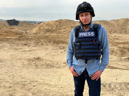 Josh Lederman stands in a desert landscape in Gaza. He is wearing a blue denim shirt, black pants, and a black bulletproof vest labeled ‘PRESS’ on the front.